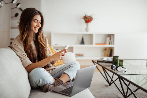 Smiling millennial mixed race woman looks at phone, reads message, has meeting, work with laptop in living room interior. App for communication remotely, study, freelance, good news at home - Foto, imagen
