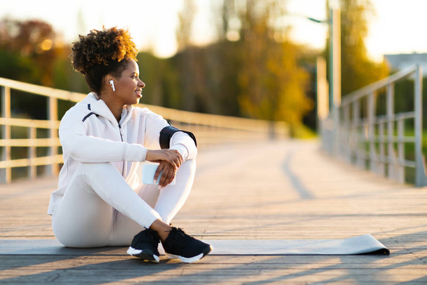 Young Black Smiling Female Sitting On Fitness Mat Outdoors, Relaxing After Training, Happy African American Woman In Sportswear Holding Smartphone And Listening Music In Wireless Earphones - Photo, Image