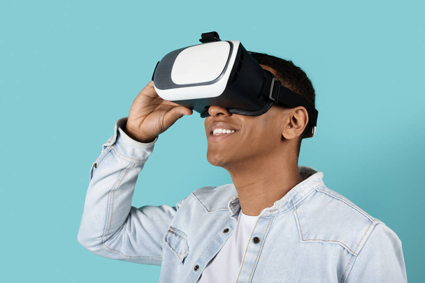 Cheerful millennial black guy in casual and vr glasses looks at blank space, enjoys online game isolated on blue studio background. Device for virtual world and fun, entertainment alone - Фото, зображення