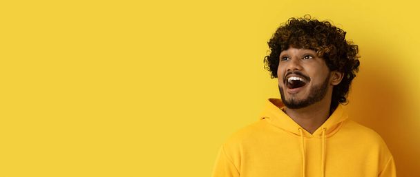Thrilled emotional amazed handsome curly young indian man in yellow hoodie looking at copy space and grimacing over yellow studio background, checking great offer or deal, web-banner - Фото, зображення