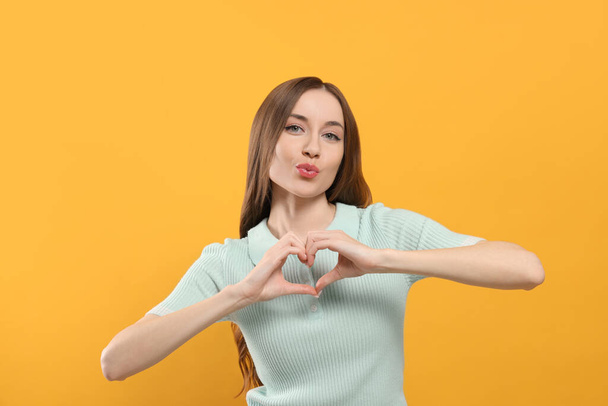Beautiful young woman blowing kiss and making heart with hands on yellow background - Foto, Imagem