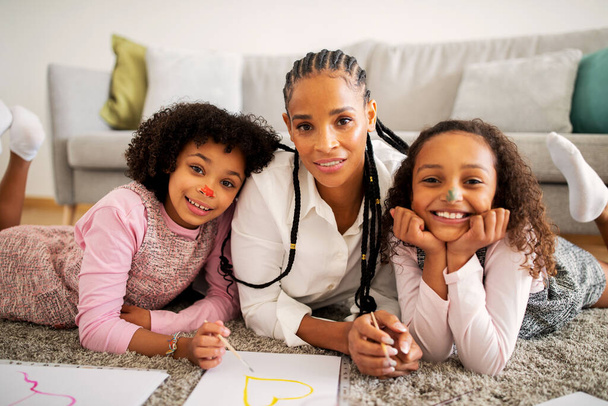 Fröhliche schwarze Mutter von zwei hübschen Töchtern beim Zeichnen und Spaß haben mit Kindern mit gemalten Gesichtern, die lächelnd vor der Kamera posieren, die zu Hause am Boden liegt. Familienwochenende Aktivität und Hobby - Foto, Bild