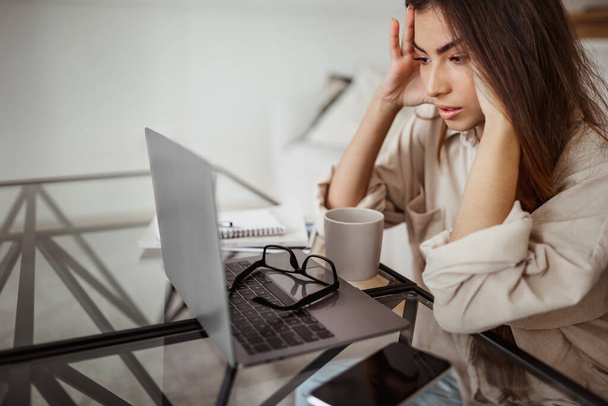 Moe ongelukkig millennial gemengde ras vrouwelijke kijk op laptop, lijden aan overwerk, druk en hoofdpijn in de kamer interieur. Problemen met werk, zaken op afstand, deadline thuis - Foto, afbeelding