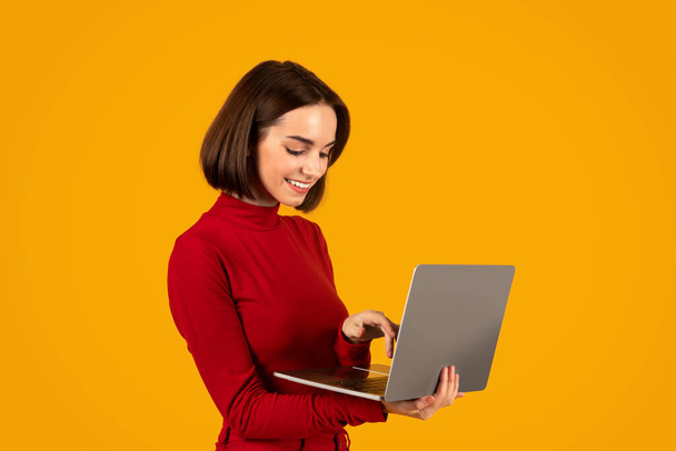 Remote job, freelance concept. Cheerful smiling pretty young woman in red with nice hairstyle typing on laptop keyboard, working remotely, isolated on orange studio background - Photo, Image