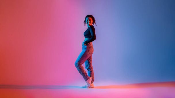 Pełna długość stylowej afro-amerykańskiej kobiety pozującej w neonowym świetle w studio i uśmiechającej się do kamery. Podekscytowana czarna dama wyrażająca radość i szczęście, pozytywne emocje koncepcja - Zdjęcie, obraz
