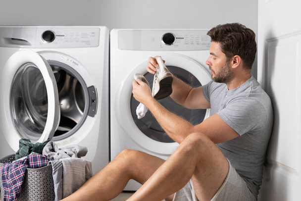 Man loading clothes into washing machine in laundry room. Middle age man putting laundry in to washing machine scared and amazed with open mouth for surprise, disbelief face with dirty clothes - Photo, image