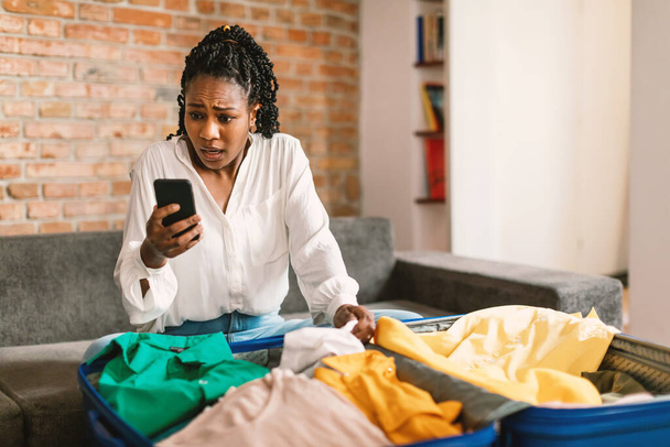 Shocked black woman reading shocking travel news about tourism on smartphone, sitting near unpacked suitcase at home. Traveling app advertisement concept - Photo, Image