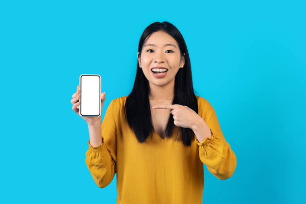 Modernes Smartphone mit weißem leeren Bildschirm in koreanischer Frauenhand, fröhliche hübsche junge Asiatin zeigt tolles Angebot oder neueste mobile App, isoliert auf blauem Studiohintergrund, Attrappe - Foto, Bild