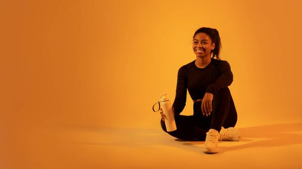 Sportif siyahi kadın elinde suyla fitness şişesi tutuyor, antrenmandan sonra dinleniyor, neon turuncu stüdyo arka planında oturuyor, boş alanı olan bir panorama, afiş. - Fotoğraf, Görsel