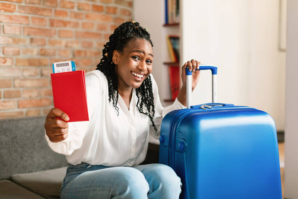 Pronti per il viaggio. Emozionata donna afroamericana in possesso di passaporto con i biglietti, seduta vicino alla valigia e sorridente alla fotocamera a casa. Vacanza, finalmente - Foto, immagini