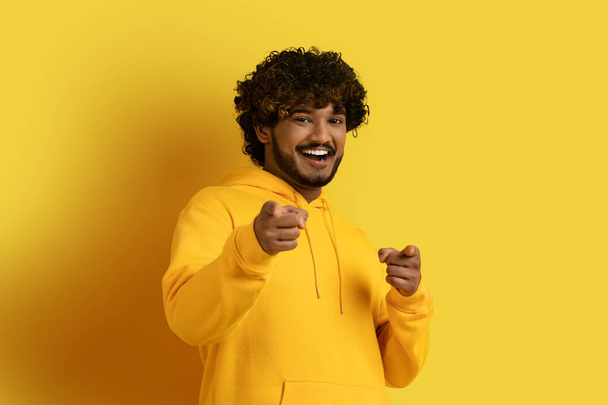 Эй ты. Счастливый игривый красивый кудрявый молодой индиец в желтой толстовке, указывающий на камеру и улыбающийся, изолированный на жёлтом фоне студии, флиртующий, наслаждающийся хорошим днем - Фото, изображение