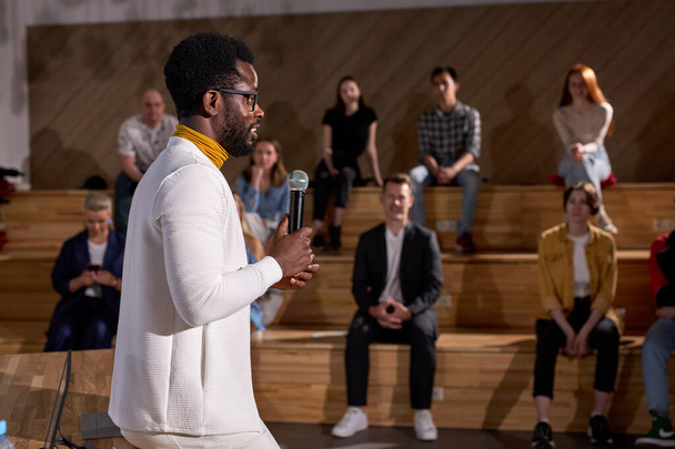 Side View Of Nice African American Teacher In White Clothing Lecturing Presenting Speech Holding Microphone In Hands, Discussing Important Theme With Students, Group Of People In The Background - 写真・画像