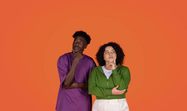 Pensive multiracial young couple african american guy and obese hispanic curly woman posing on red studio background together, touching chin and looking at copy space, thinking about something - Photo, image