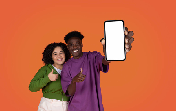 ショッピングモバイルアプリ、オンラインオファー、電子商取引。幸せな陽気な美しい多人種の若いカップルが白い空の画面と親指のアップ、赤いスタジオの背景、モックアップ、コピースペースとスマートフォンを示しています - 写真・画像