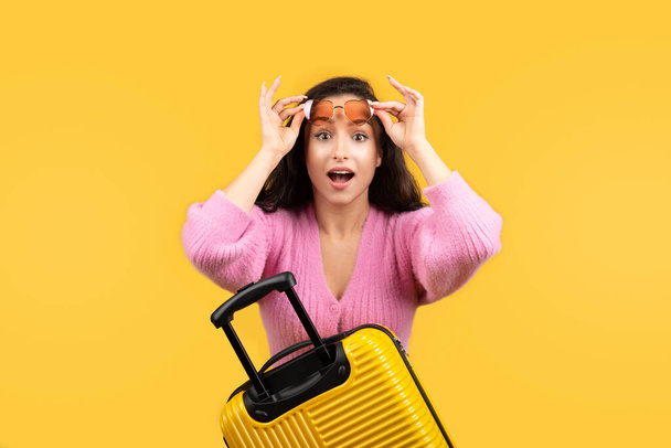 Cheerful shocked excited young caucasian brunette woman takes off glasses, enjoy trip with suitcase isolated on yellow studio background. Travel, tourism, vacation and great offer - 写真・画像