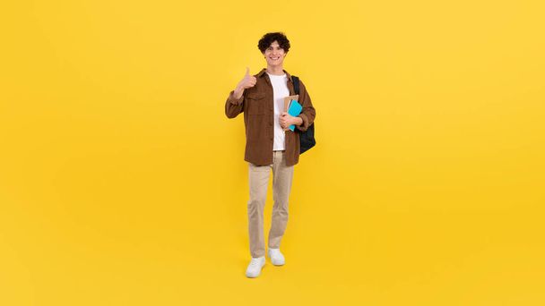 Happy Student Guy Gesturing Thumbs Up Approving University Educational Program Posing With Backpack Holding Notebooks Standing Over Yellow Studio Background, Smiling To Camera. Panorama - Photo, Image