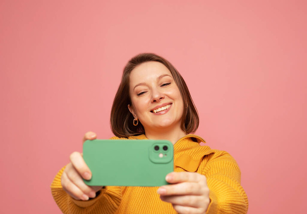 Cheerful funny woman 40s wearing yellow sweater posing doing selfie shot on mobile phone over pink background - Фото, зображення