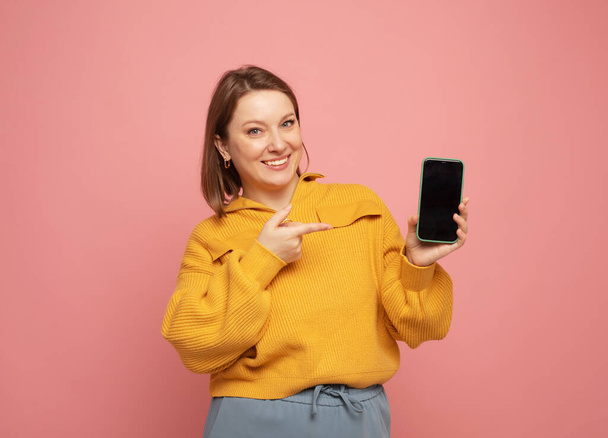 smiling adult woman wearing yellow sweater showing or presenting mobile phone over pink background - Photo, image
