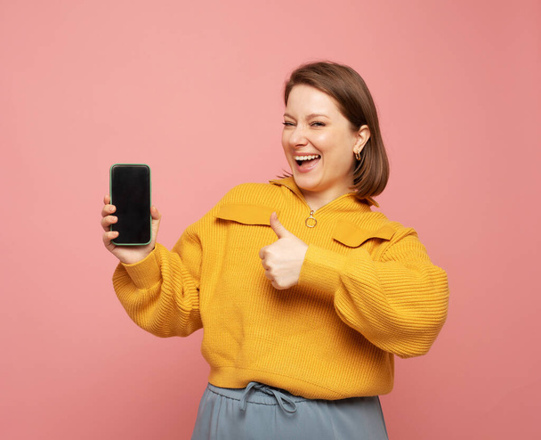 mosolygós felnőtt nő visel sárga pulóver bemutató vagy bemutató mobiltelefon rózsaszín háttér - Fotó, kép