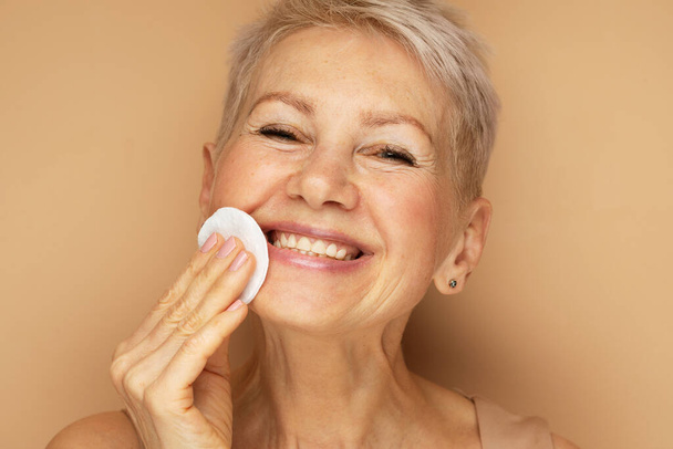 Mature woman cleansing her face with cotton pad looking at camera. Beauty and skincare routine - Foto, Imagen