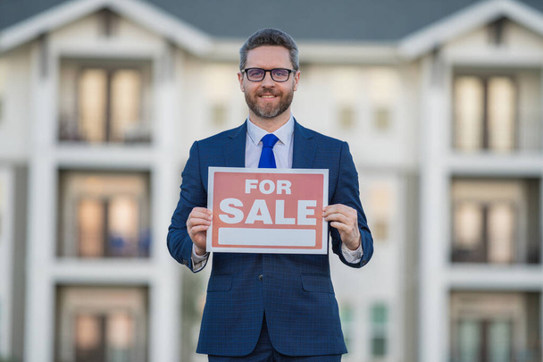 Successful real estate agent in a suit holding for sale sign near new apartment. Real estate agent with home loan contract, selling home. Realtor or real estate agent shows board for sale - 写真・画像