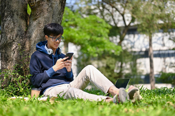 Щасливий і гарний молодий азійський студент коледжу, який користується своїм смартфоном, розмовляє з друзями або грає в мобільні ігри, сидячи під деревом у парку.. - Фото, зображення