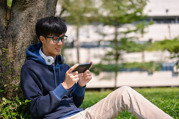 Portret van een gelukkige jonge Aziatische student die zijn smartphone gebruikt of mobiele spelletjes speelt terwijl hij onder de boom zit in het campuspark. - Foto, afbeelding