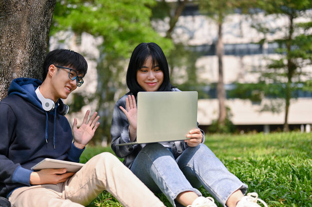 Two smiling and happy young Asian college students are having an online meeting or online class with their teacher while sitting on the grass at the campus park. - Foto, Bild