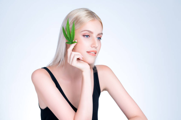 Kellemes gyönyörű szőke haj tökéletes sima smink bőr tartsa cannabis zöld kender elszigetelt háttérben a természetes CBD bőrápoló kezelés expresszív arc és gesztus kifejezés. - Fotó, kép