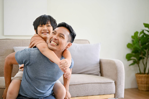 喜びと幸せなアジアのお父さんは、リビングルームで彼の小さな息子と遊んでいます,ピギーバック,飛行機をプレイ,一緒に家で楽しい時間を過ごす. - 写真・画像