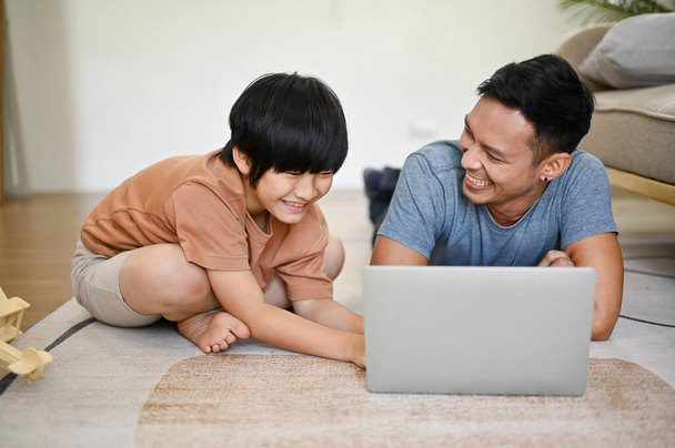 Грайливий і щасливий азіатський тато лежить на підлозі, спостерігаючи фільм на ноутбуці у вітальні зі своїм сином на вихідних. - Фото, зображення