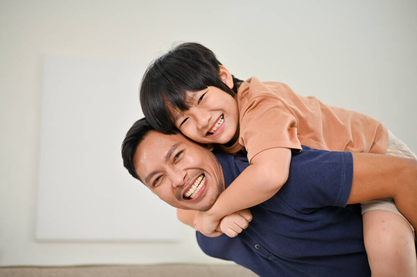Mutlu Asyalı oğul ve baba evde birlikte eğleniyor, oturma odasında oynuyorlar ve sırtımda taşıyorlar. Mutlu aile ve bekar baba kavramı - Fotoğraf, Görsel