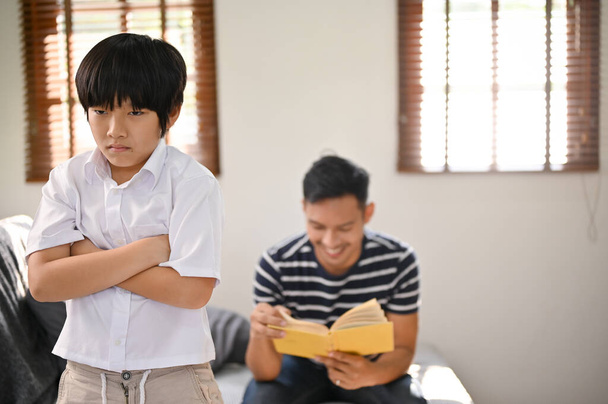 Kızgın ve üzgün bir Asyalı çocuk, kollarını salonda kavuşturmuş, yalnız ve ihmal edilmiş hissediyor, arkasında kitap okuyan babası tarafından.. - Fotoğraf, Görsel