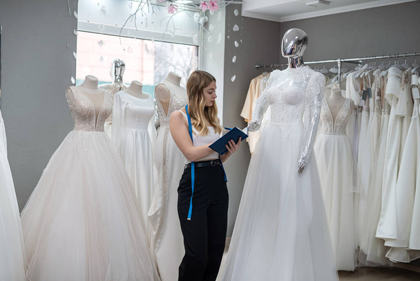 Pretty  female dressmaker with diary and pen posing near bridal wear in wedding fashion boutique - 写真・画像