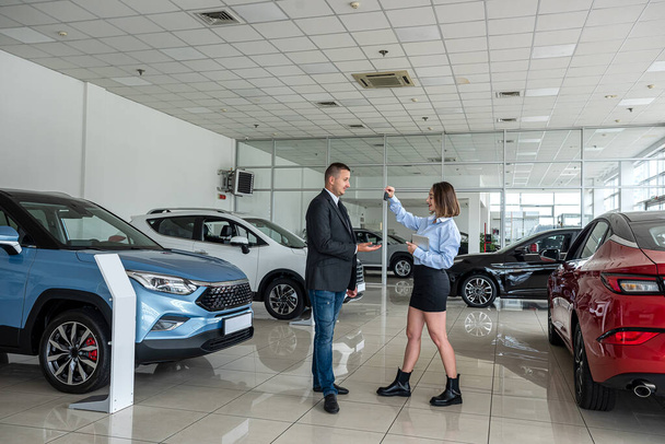 Young pretty female salesmoman giving a key to young happy man client in car showroom. sale or rent concept - Φωτογραφία, εικόνα