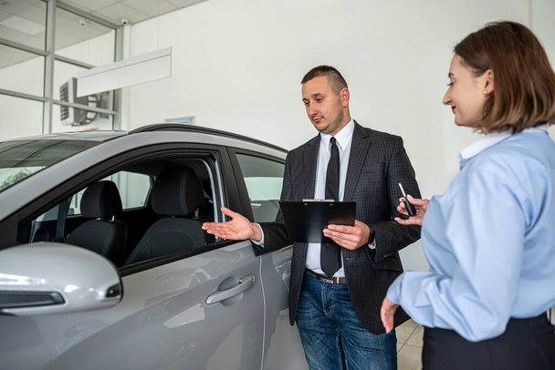  Galeride modern bir arabanın yanında duran genç ve güzel bir bayan satıcıdan yardım alarak en iyi otomobili seçiyor. satın alma kavramı - Fotoğraf, Görsel