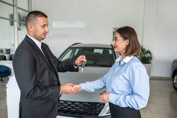 Jonge mooie vrouwelijke salesmoman geeft een sleutel aan jonge gelukkige man klant in de auto showroom. verkoop- of huurconcept - Foto, afbeelding