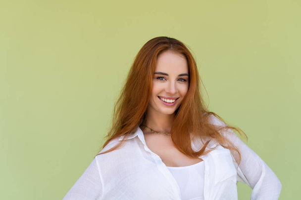 Yeşil arka planda rahat gömlekli, uzun kızıl saçlı güzel bir kadın gülümsüyor ve eğleniyor. - Fotoğraf, Görsel