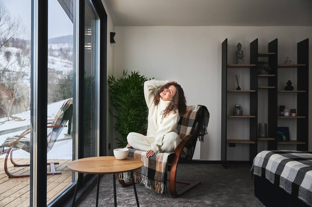 Curly young woman enjoying weekends inside contemporary barn house in the mountains. Happy female tourist sitting on chair, holding cup tea in new cottage with panoramic windows. - Фото, изображение