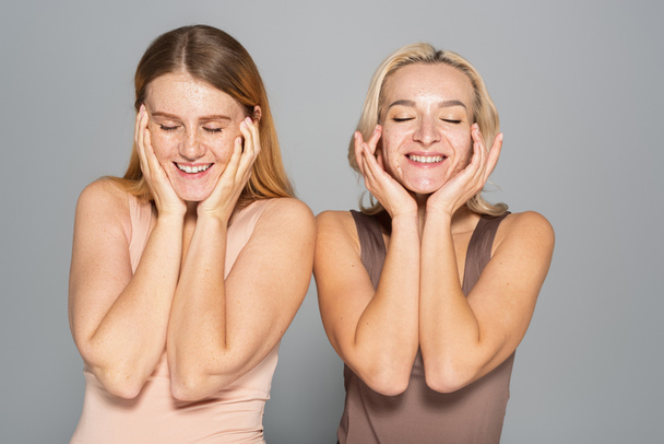 Uśmiechnięte, międzyrasowe kobiety z problematyczną skórą dotykające twarzy odizolowane na szaro  - Zdjęcie, obraz
