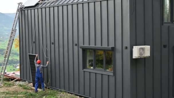 Erkek işçiler evin çatısına güneş paneli kuruyor. Kasklı elektrikçiler fotovoltaik güneş modülünü açık havada halatların yardımıyla kaldırıyorlar. Alternatif ve yenilenebilir enerji kavramı. - Video, Çekim