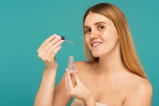 Smiling woman with freckled skin holding serum and dropper isolated on turquoise - Fotó, kép