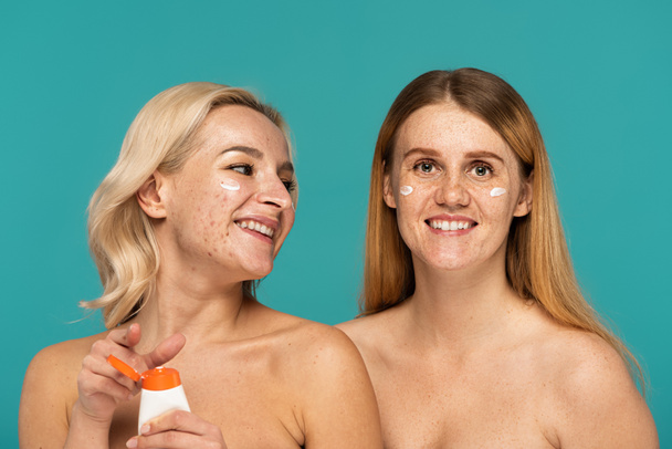 giovani donne felici con diverse condizioni della pelle che applicano la crema sui volti isolati sul turchese  - Foto, immagini