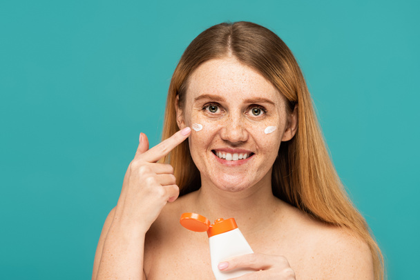 mujer feliz con pecas sosteniendo tubo y aplicando crema cosmética aislada en turquesa - Foto, imagen
