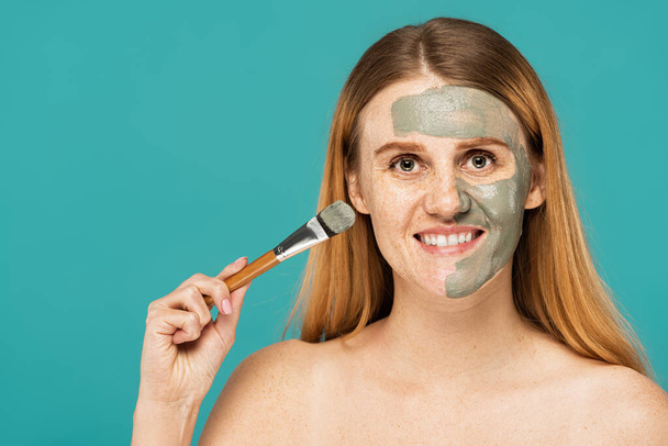 veselá žena s rudými vlasy drží kosmetický kartáč při aplikaci hliněné masky na polovinu obličeje izolované na tyrkysové  - Fotografie, Obrázek