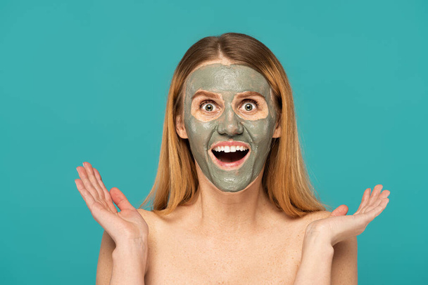 verbaasd vrouw met rood haar en klei masker op gezicht gebaar geïsoleerd op turquoise - Foto, afbeelding