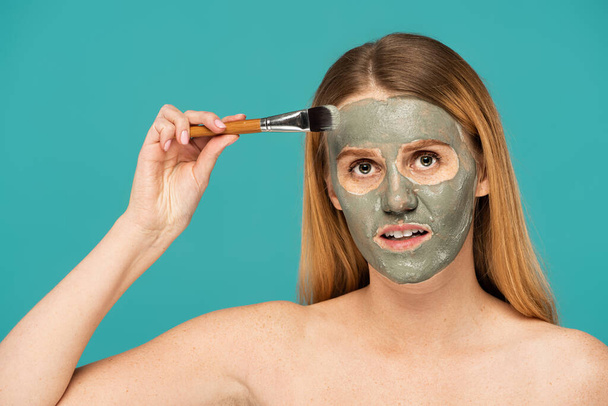 young woman with red hair applying clay mask on face isolated on turquoise - Foto, immagini