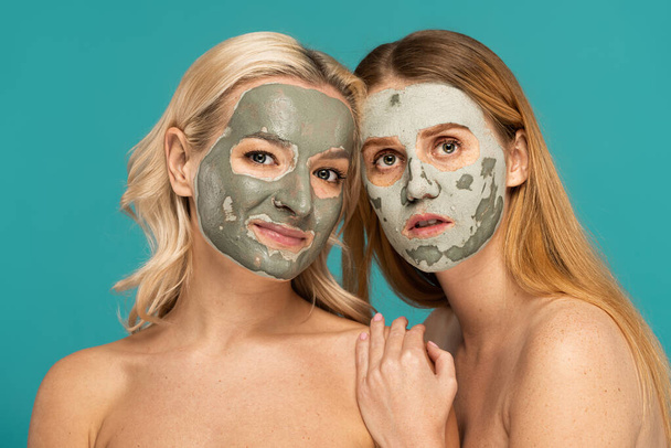 zrzky a blondýny ženy s hliněnou maskou na tvářích při pohledu na kameru izolované na tyrkysové - Fotografie, Obrázek
