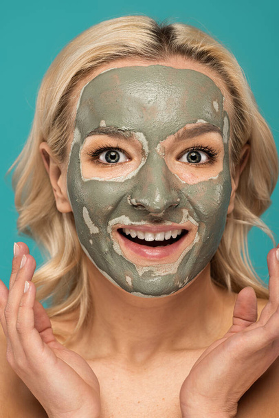opgewonden blonde vrouw met klei masker op gezicht kijken naar camera geïsoleerd op turquoise - Foto, afbeelding