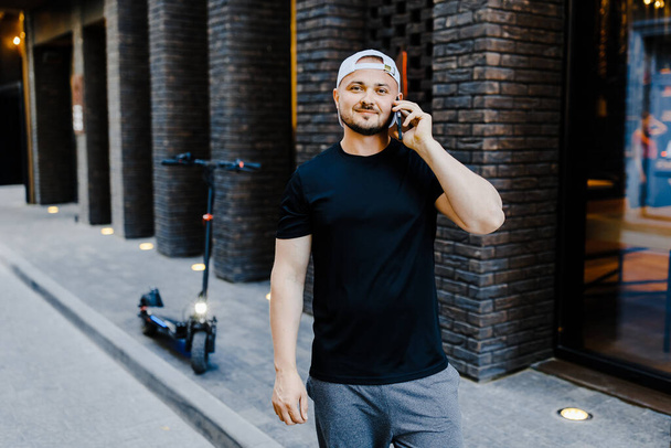 Valkoihoinen nuori komea mies seisoo kadulla lähellä sähköskootteria ja puhuu matkapuhelimella. - Valokuva, kuva